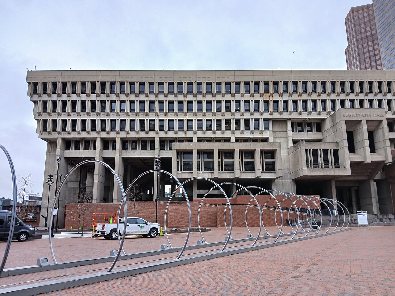 A large concrete building with deep overhanging upper floors stands above a brick plaza with a row of metal arches.