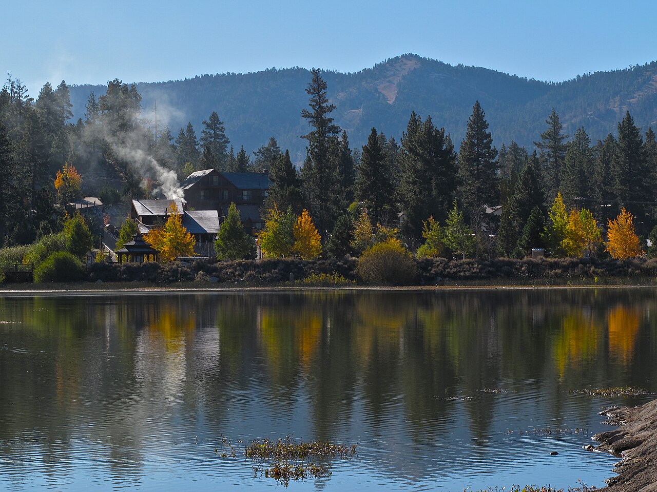 A lakeside house with a smoking chimney sits among pines and yellow autumn trees, all mirrored in still water.