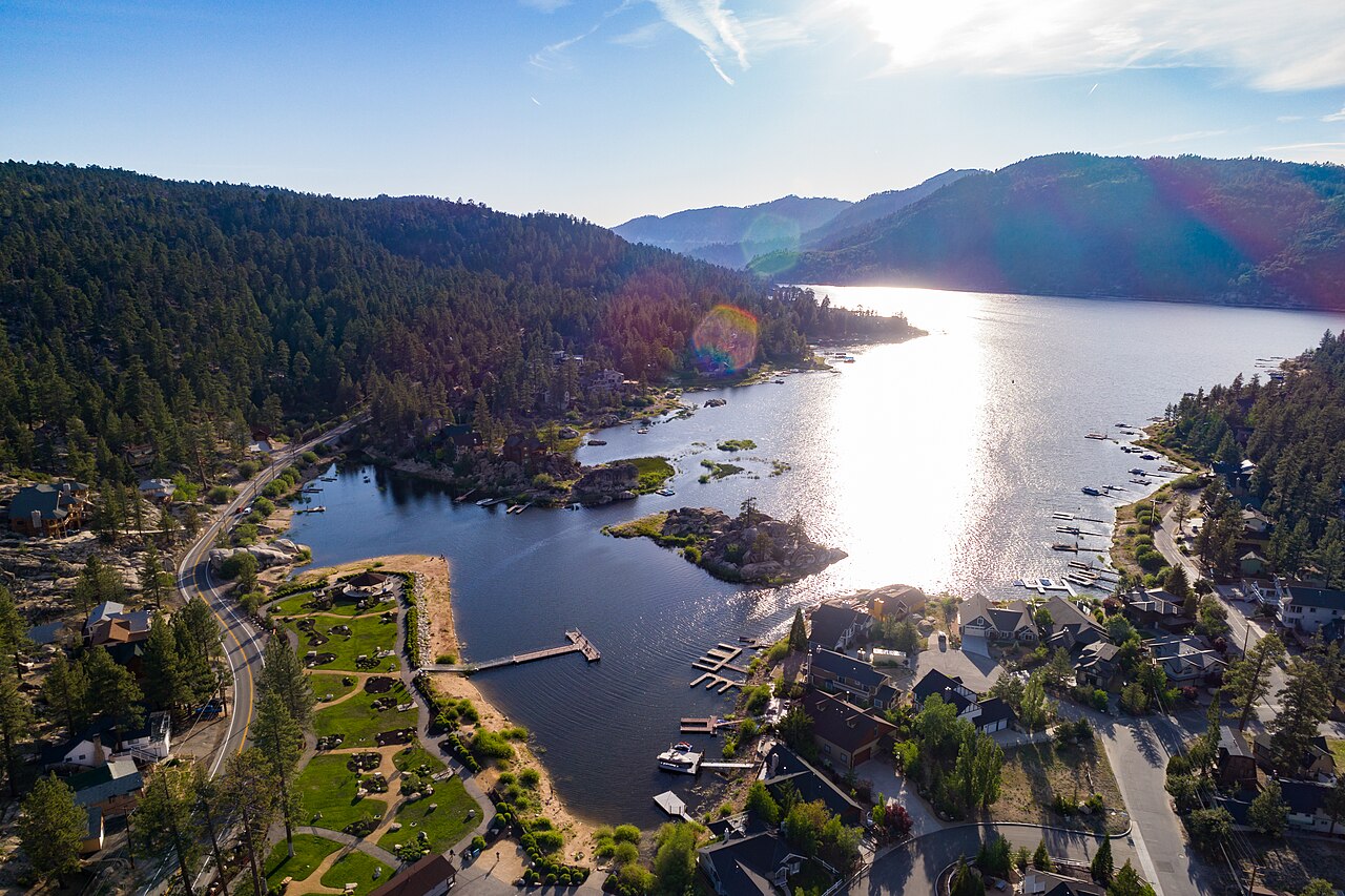 An aerial view of a mountain lake with small docks, a shoreline road and houses, with sunlight glaring off the water.