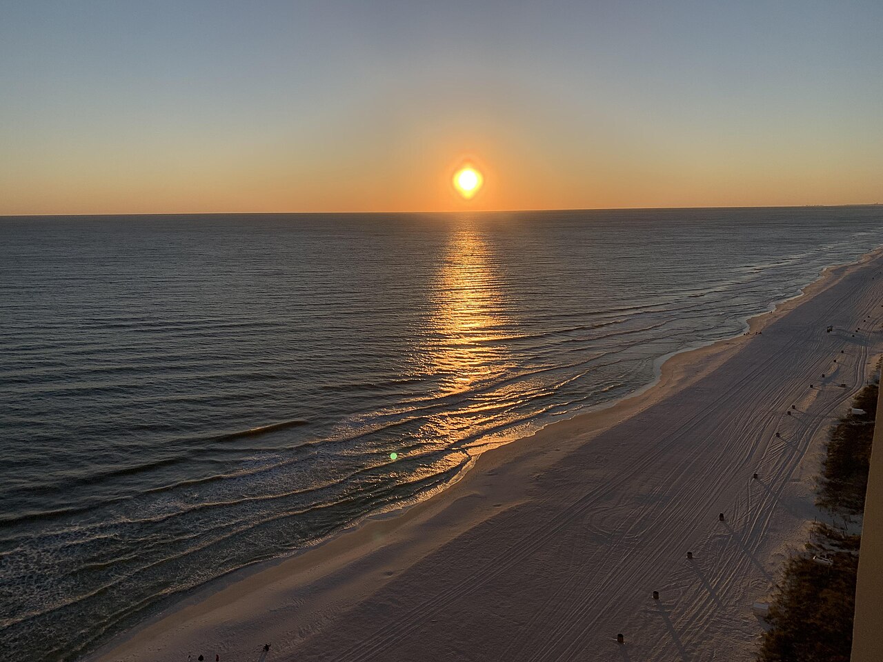 A low sun hangs over a calm sea, laying a strip of orange light across the water toward a curving sandy shore.