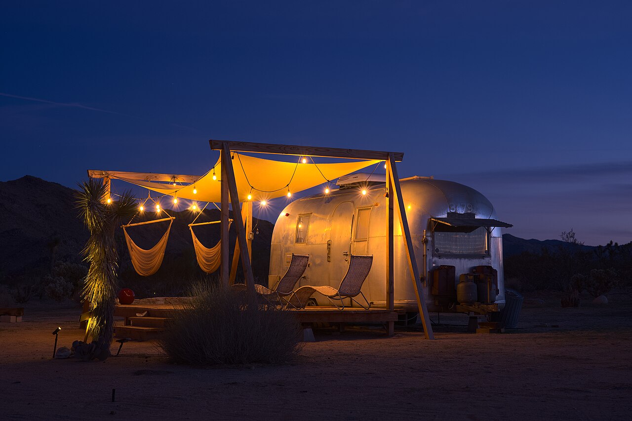 A polished aluminium trailer under a lit canvas canopy with hammocks and string lights, beneath a deep blue evening sky.