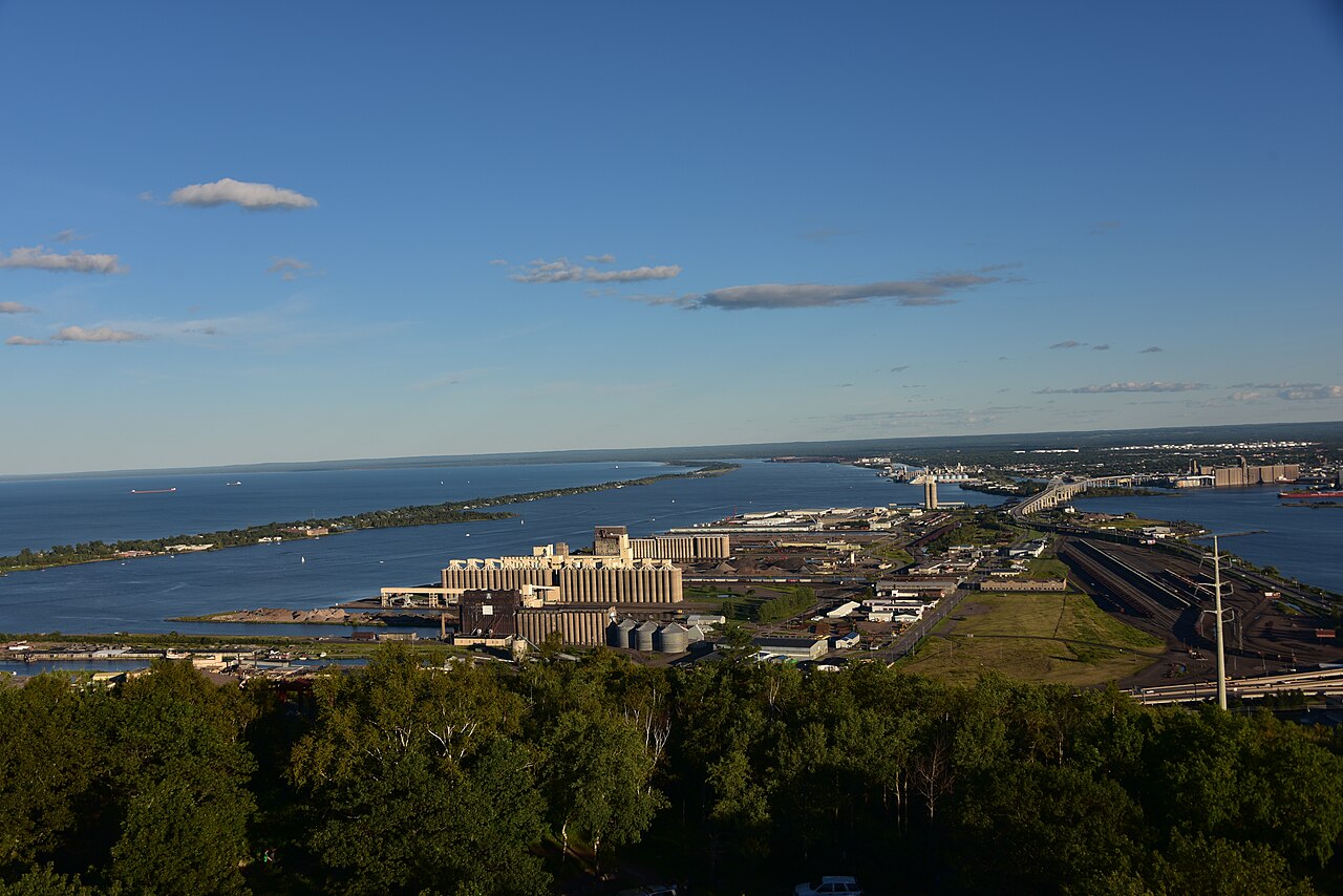 A high view over treetops to a long strip of land with docks and industrial buildings reaching into a wide blue harbour.