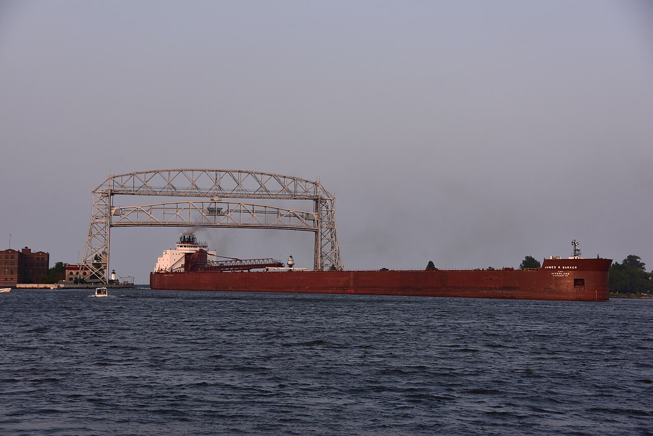 A long red cargo ship passes beneath a raised steel lift bridge on grey water under an overcast sky.