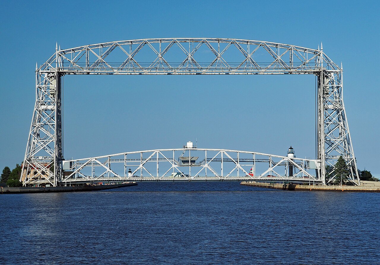 A steel lift bridge with two tall towers spans a canal, with blue water in front and a clear sky behind.