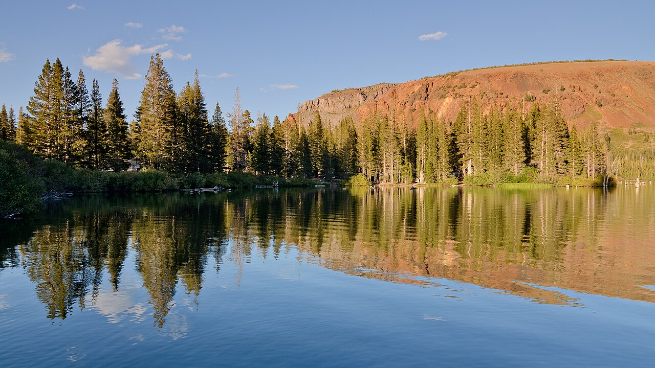 A still mountain lake reflects tall pine trees and a red-brown ridge under a clear sky.