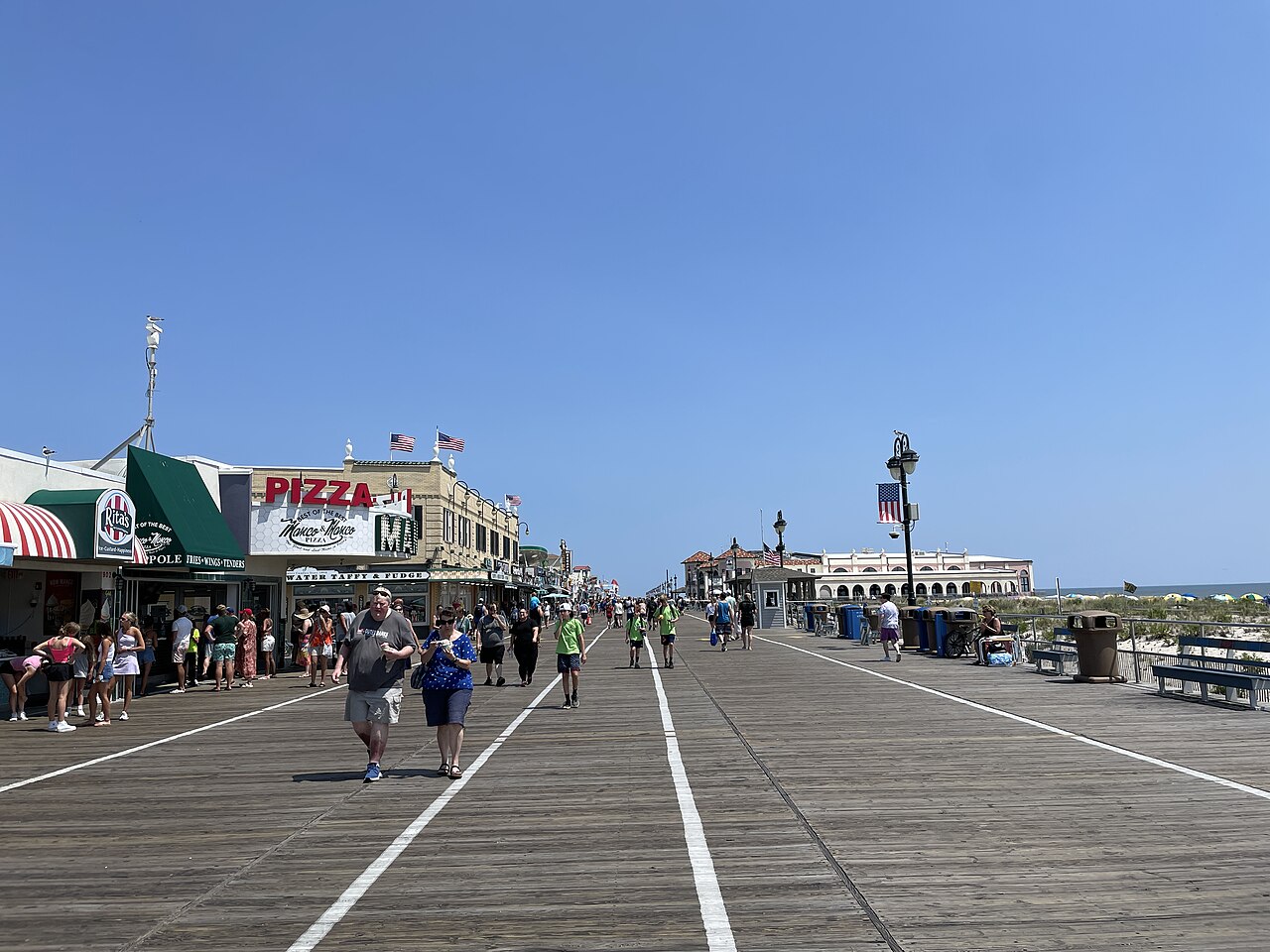 A broad wooden boardwalk lined with shops and flags runs into the distance under a clear blue sky, with people walking.