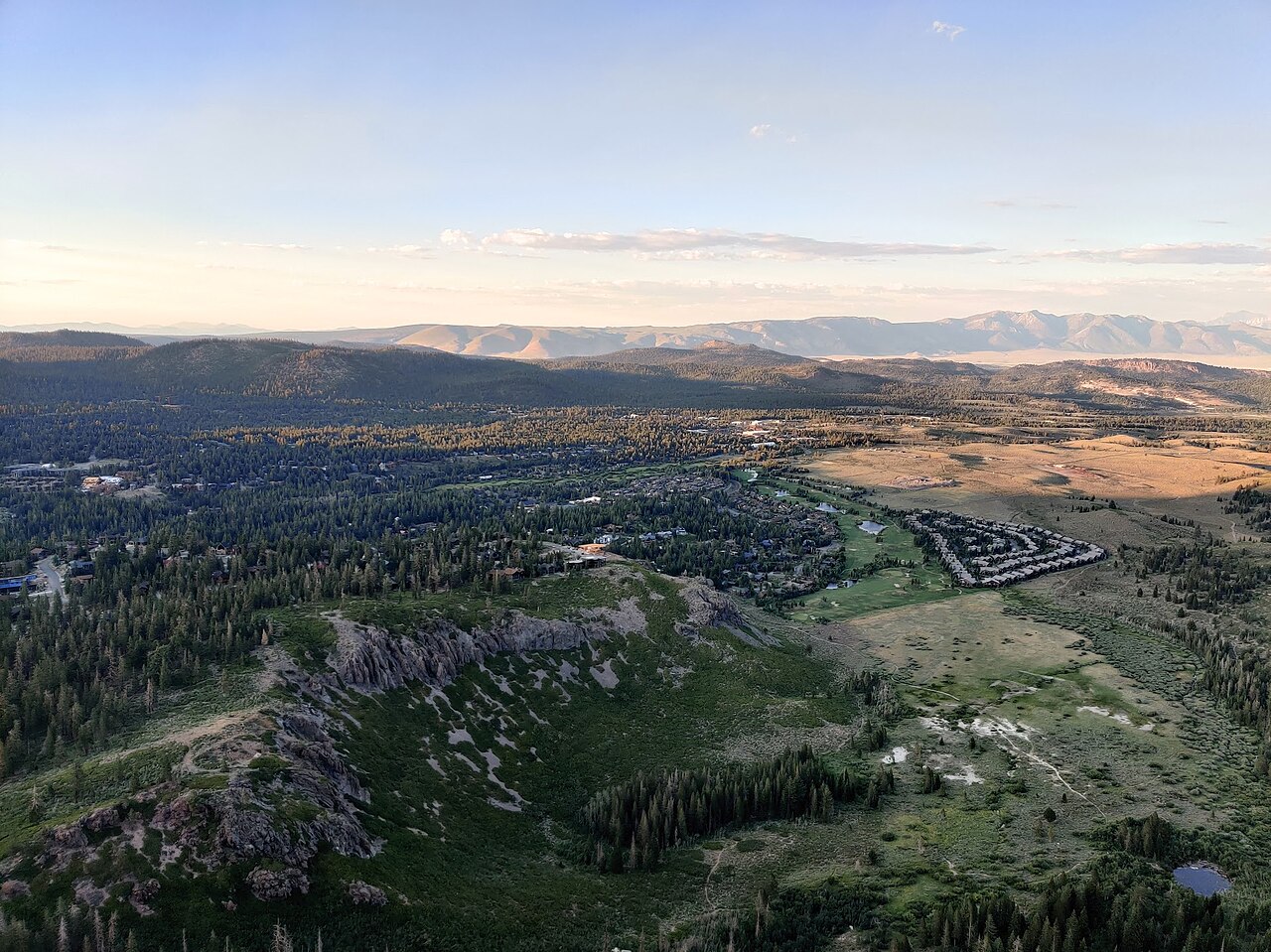 A wide view from above of a small town spread among pine forest, with dry hills and distant mountains in evening light.