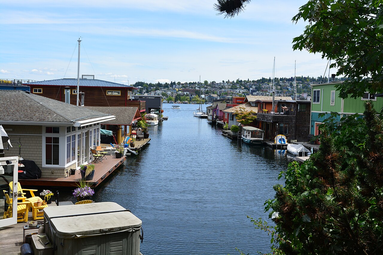 Two rows of low floating houses face each other across a channel of calm water that opens onto a lake and a wooded hillside.