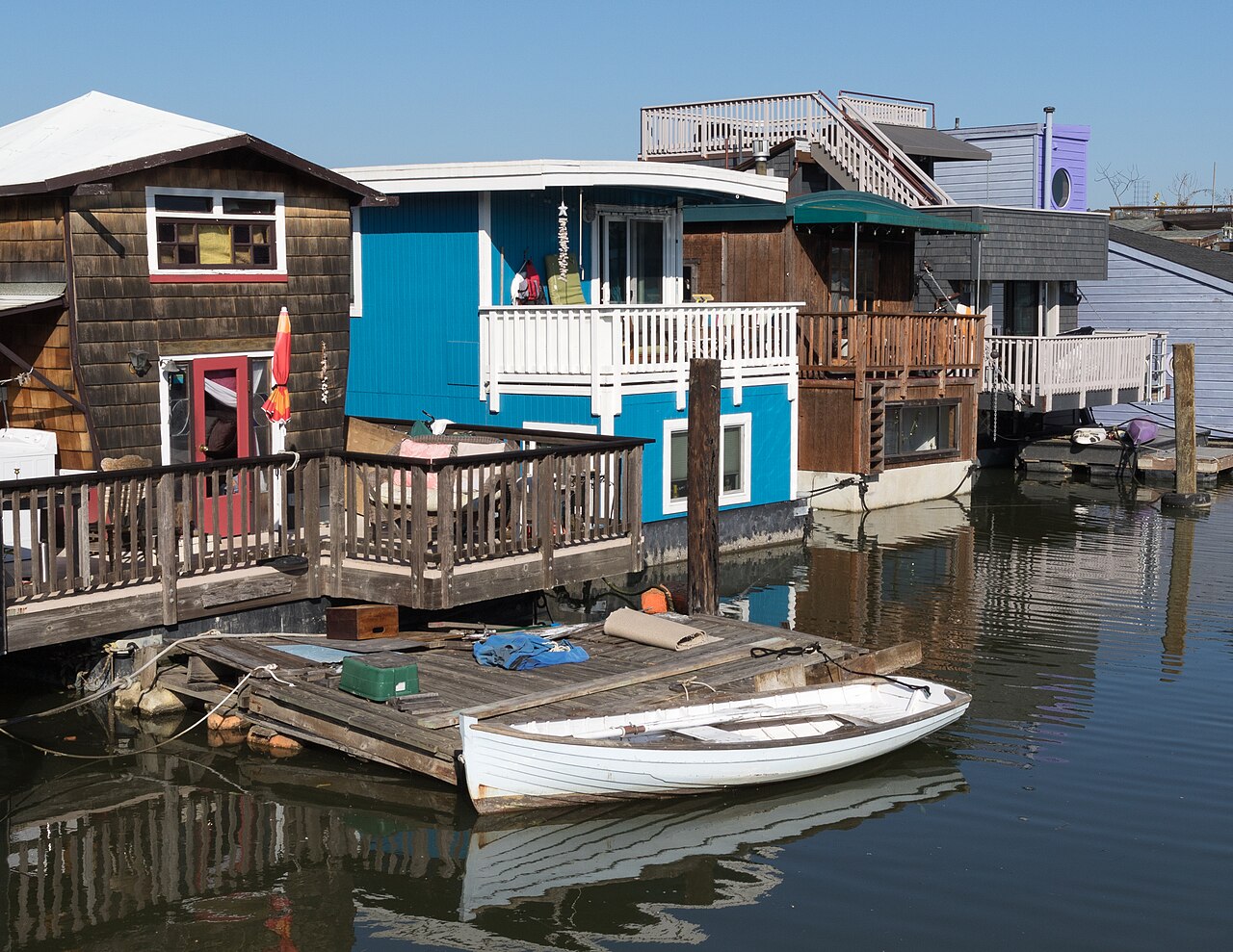 Wooden houseboats painted brown and bright blue sit side by side on the water, with a white rowing boat tied to a small dock.