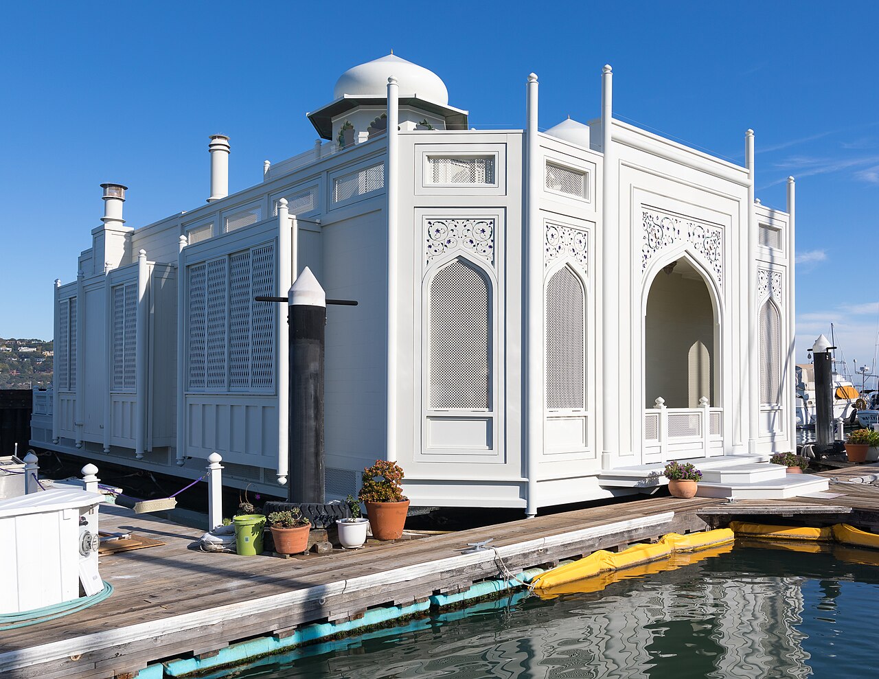 A white floating house with pointed arches, carved screens and a small dome sits beside a wooden dock under a deep blue sky.