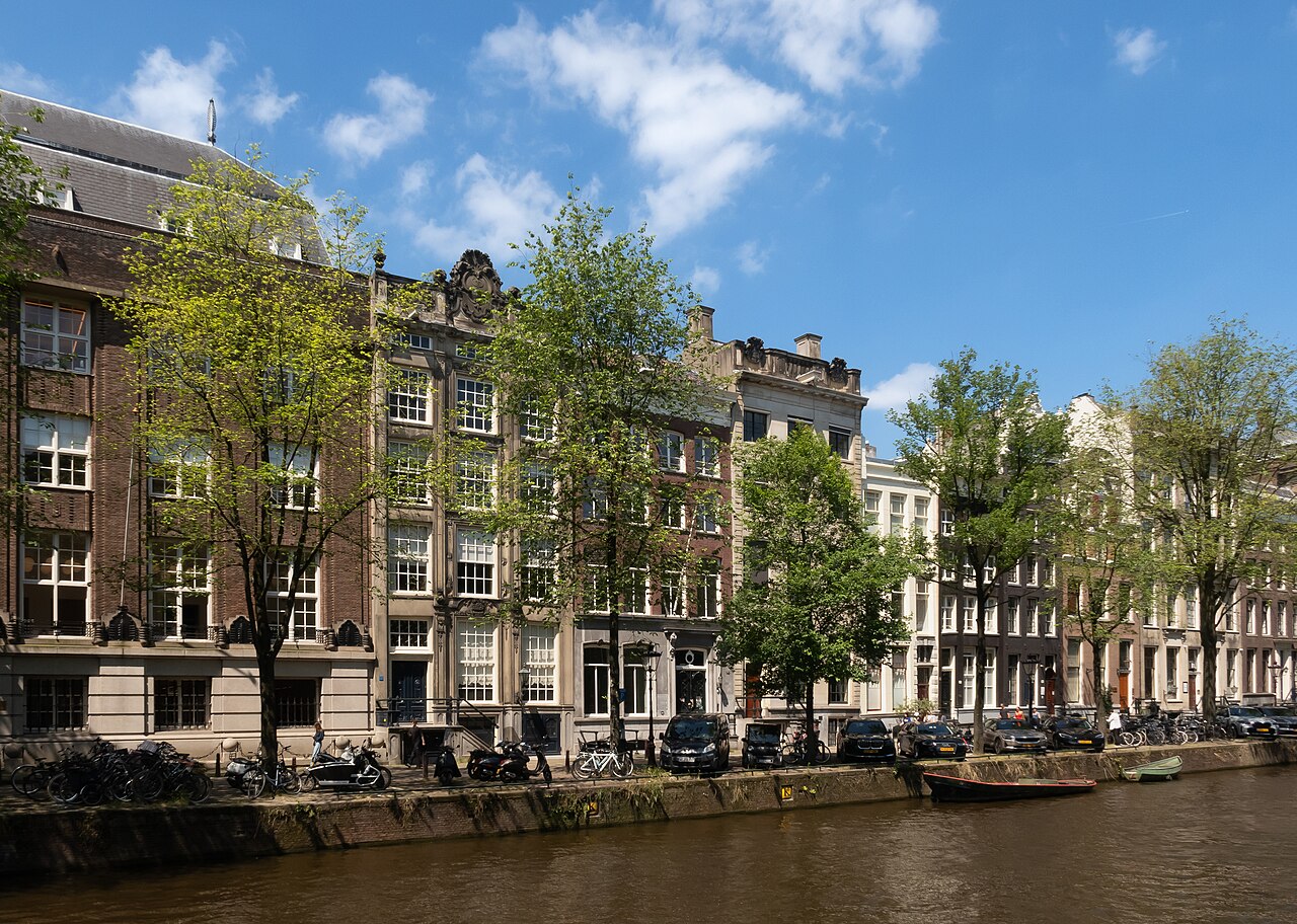 A row of tall narrow brick houses with gabled tops stands behind trees along a canal with moored boats.