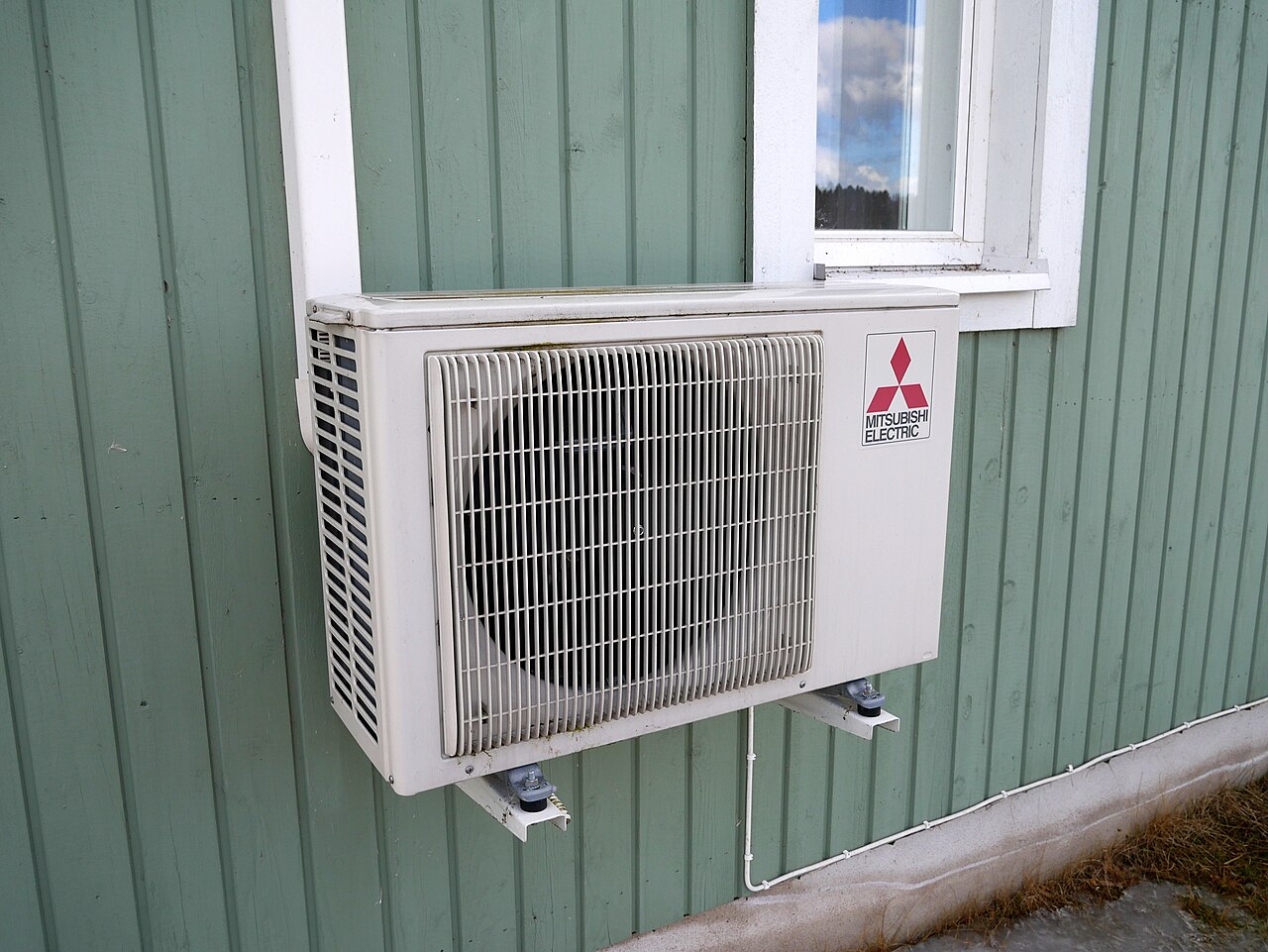 A white box-shaped unit with a round fan grille is fixed on brackets to a green timber-clad wall below a window.