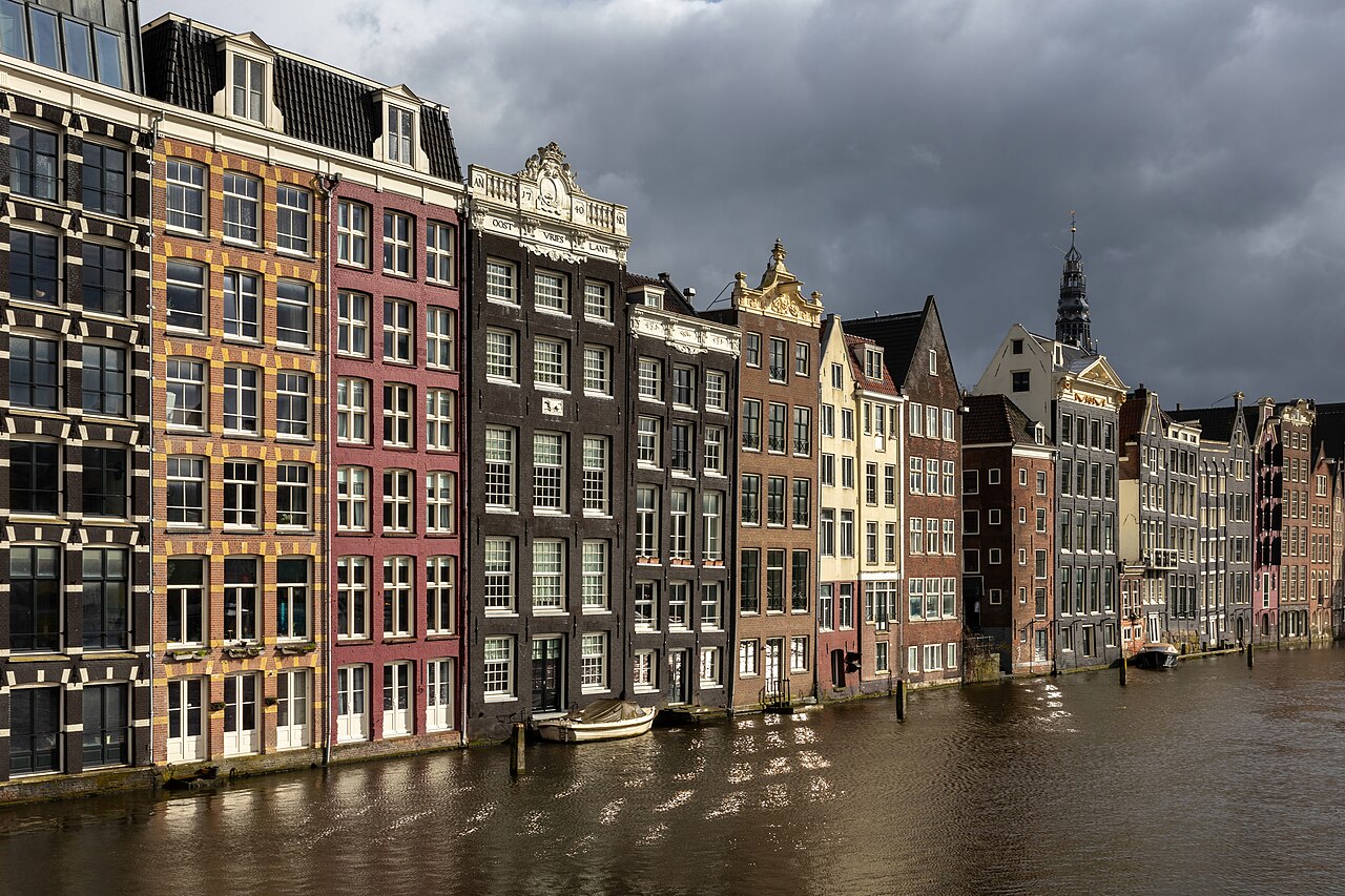 A row of tall narrow houses in red, brown and cream rises straight out of the water under a dark stormy sky.