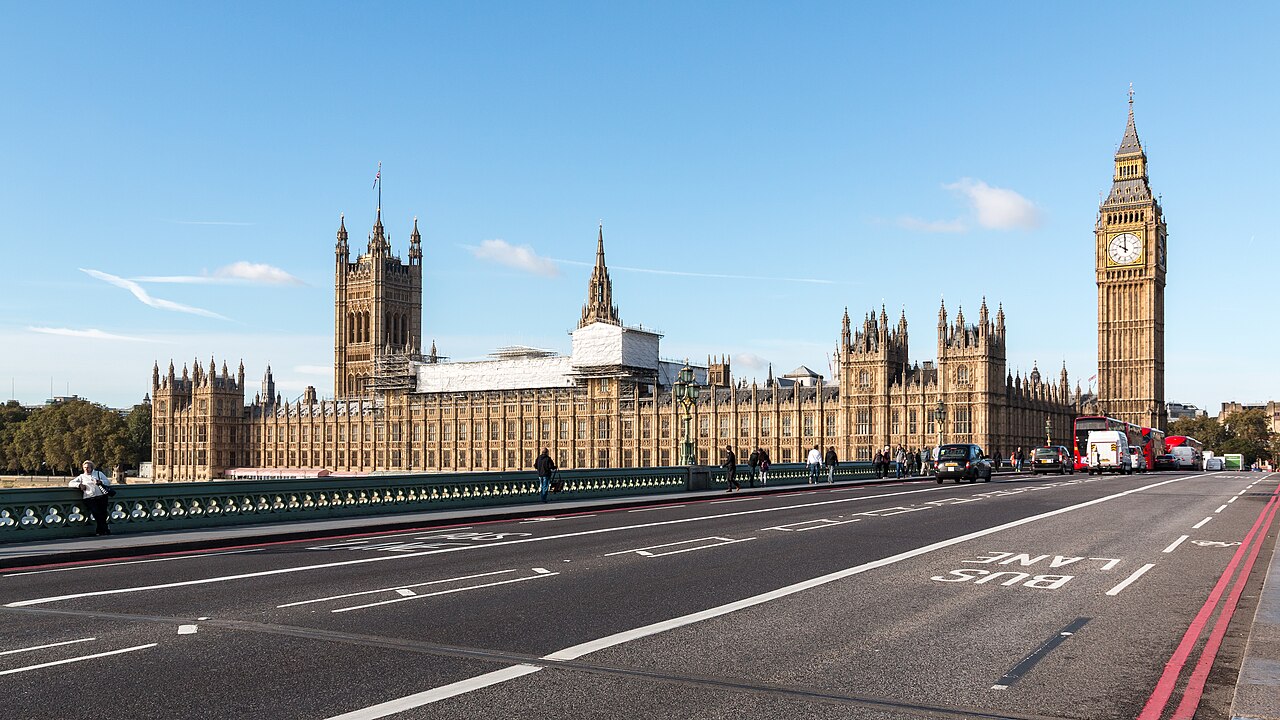 A long Gothic stone building with towers and a tall clock tower stands beyond an empty bridge roadway under a blue sky.