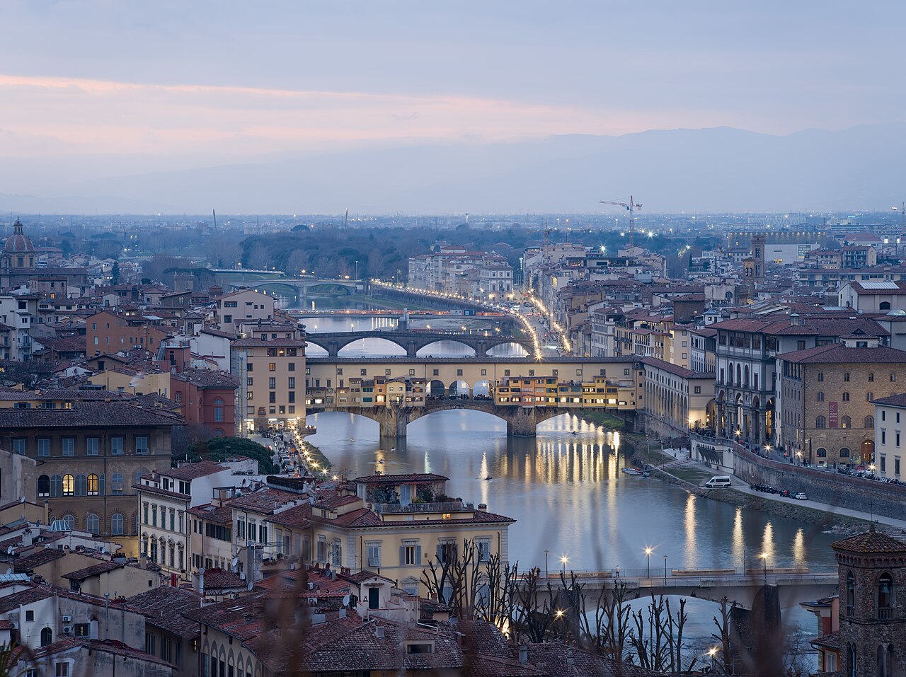 A river crossed by several stone bridges runs through a dense city of tiled roofs as the lights come on at dusk.