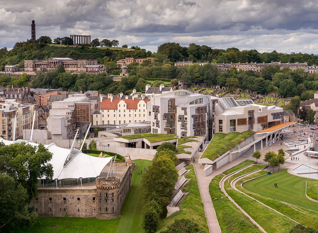 A cluster of low modern buildings with curved roofs sits among green landscaped terraces, with a wooded hill behind.