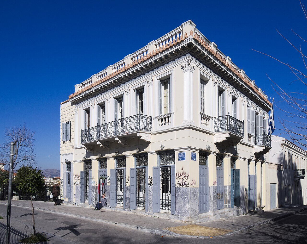 A white two-storey corner house with iron balconies and a decorated roofline stands against a deep blue sky.