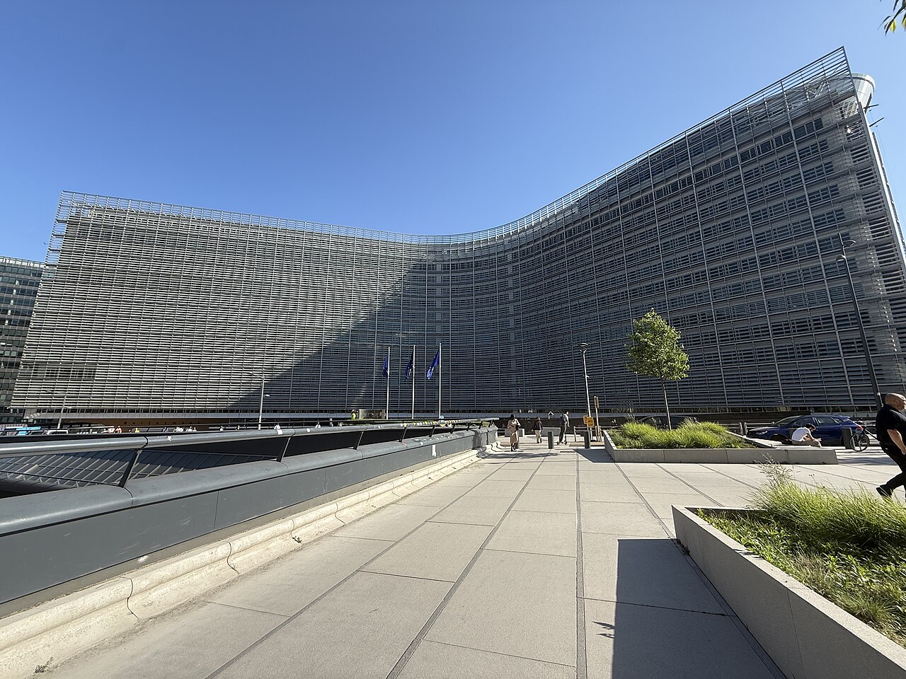 A large curved office building with a dark glass facade rises behind a paved forecourt with planters under a blue sky.