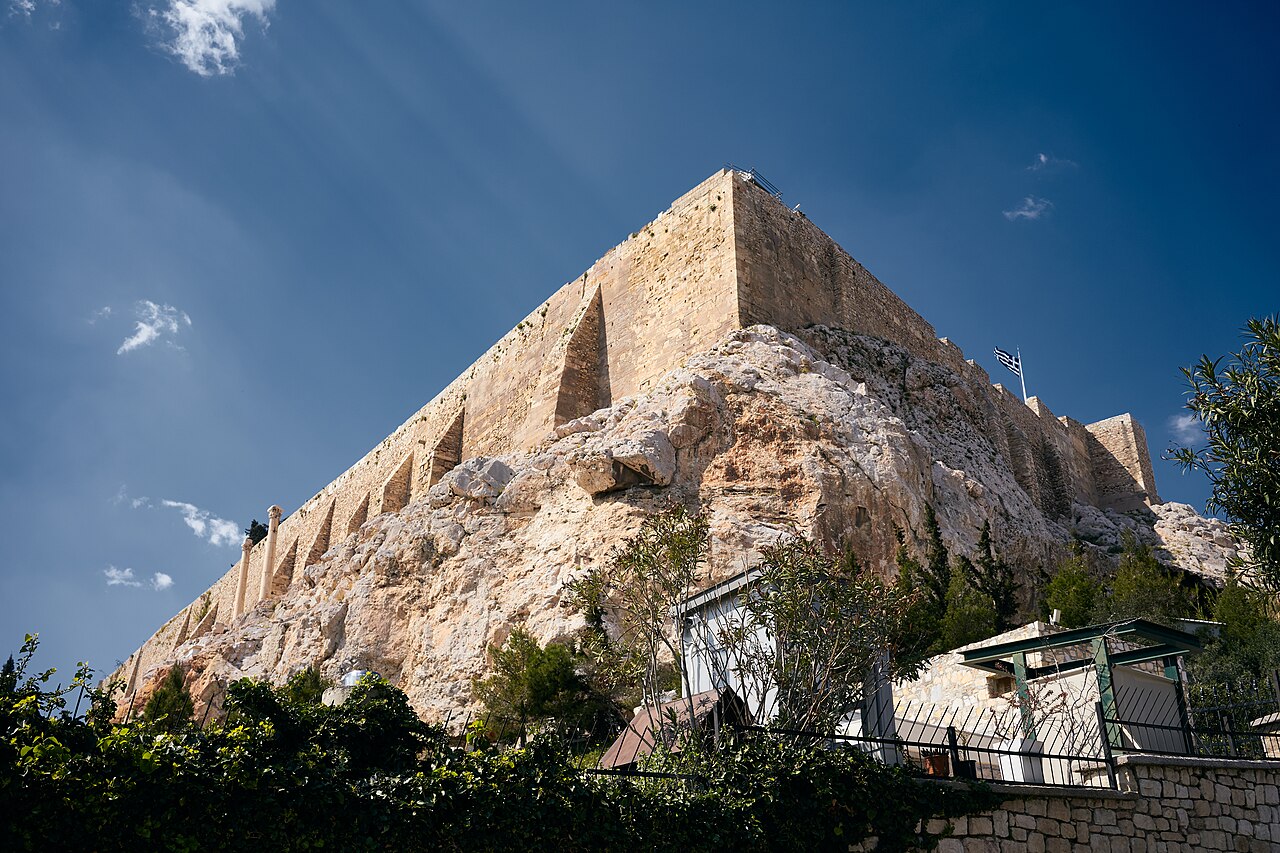 A steep rock topped by high ancient stone walls rises above trees and rooftops under a blue sky.