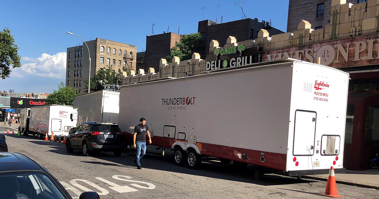 Long white production trailers line the kerb of a residential street of brick apartment buildings.