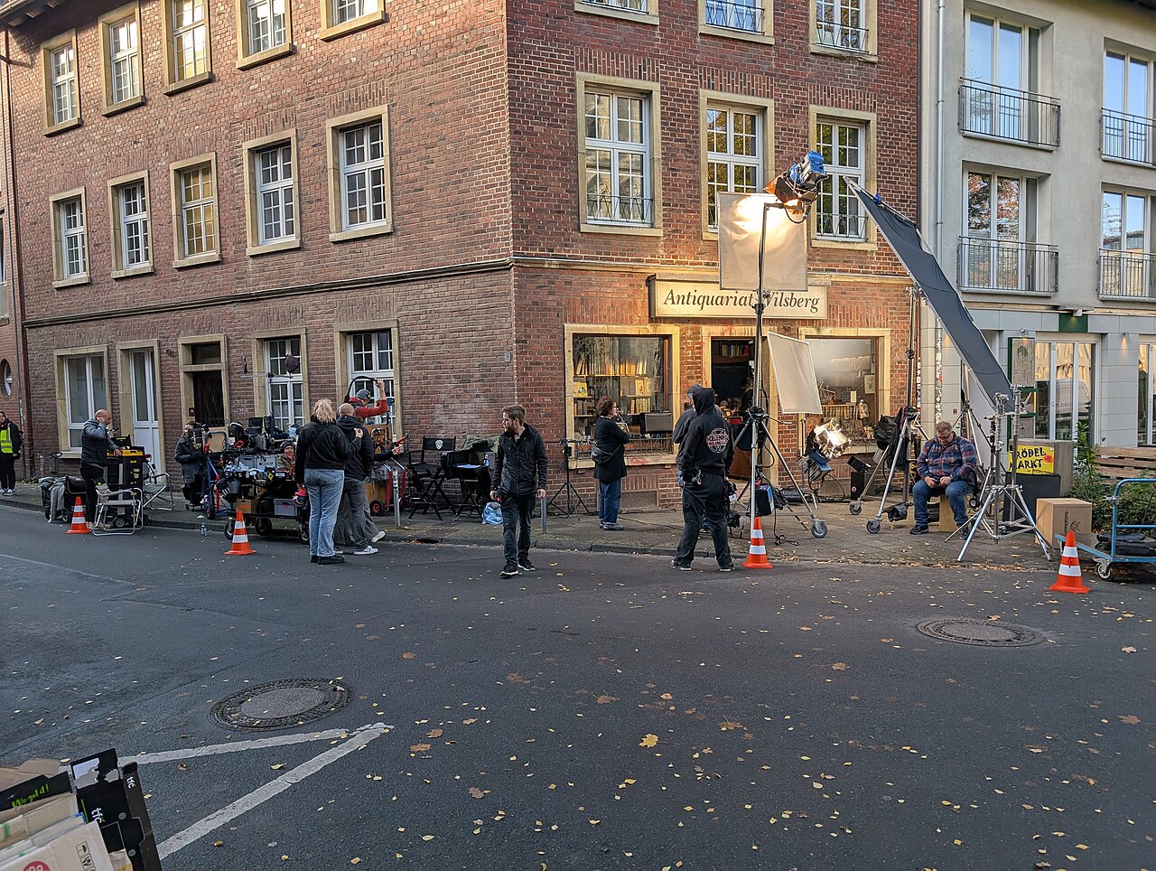 A group of crew members stands with lamps, reflector screens and stands outside a brick corner building on a city street.