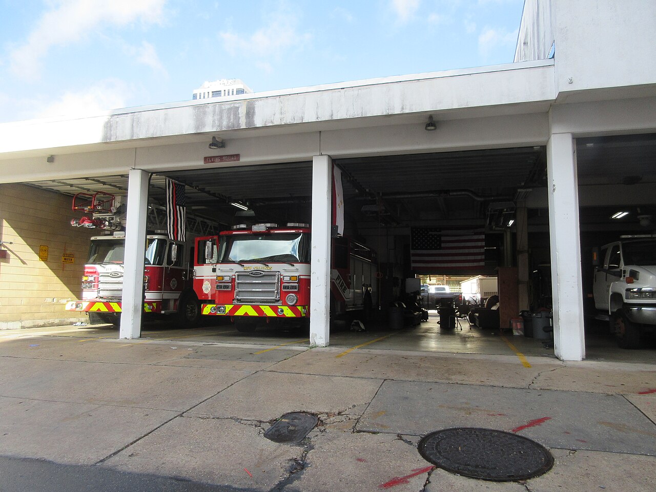 Red and white fire engines stand inside the open bays of a low white fire station with a wide concrete forecourt.