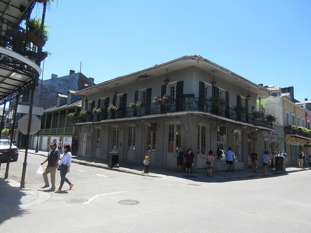A two-storey corner building with a long iron balcony full of plants stands at a quiet street crossing.