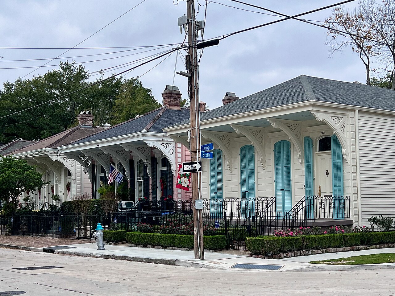 A row of single-storey wooden houses with decorated porches and turquoise shutters stands behind an iron fence on a street corner.