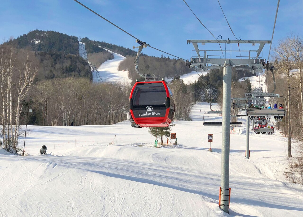 A red gondola cabin hangs from a lift cable above a snowy slope, with lift towers and bare trees behind.