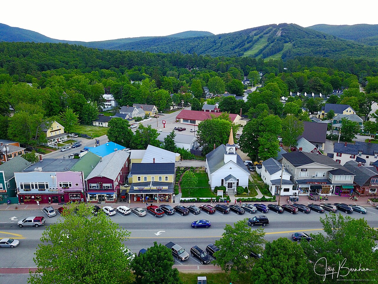 A village main street lined with shops and parked cars, a white church among trees, and a green mountain with ski trails behind.
