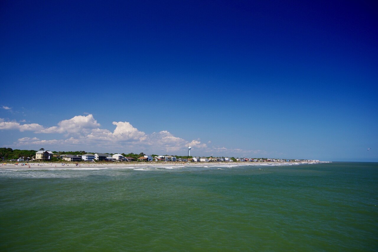 Green ocean water in the foreground with a low shoreline of beach houses on the horizon under a deep blue sky.