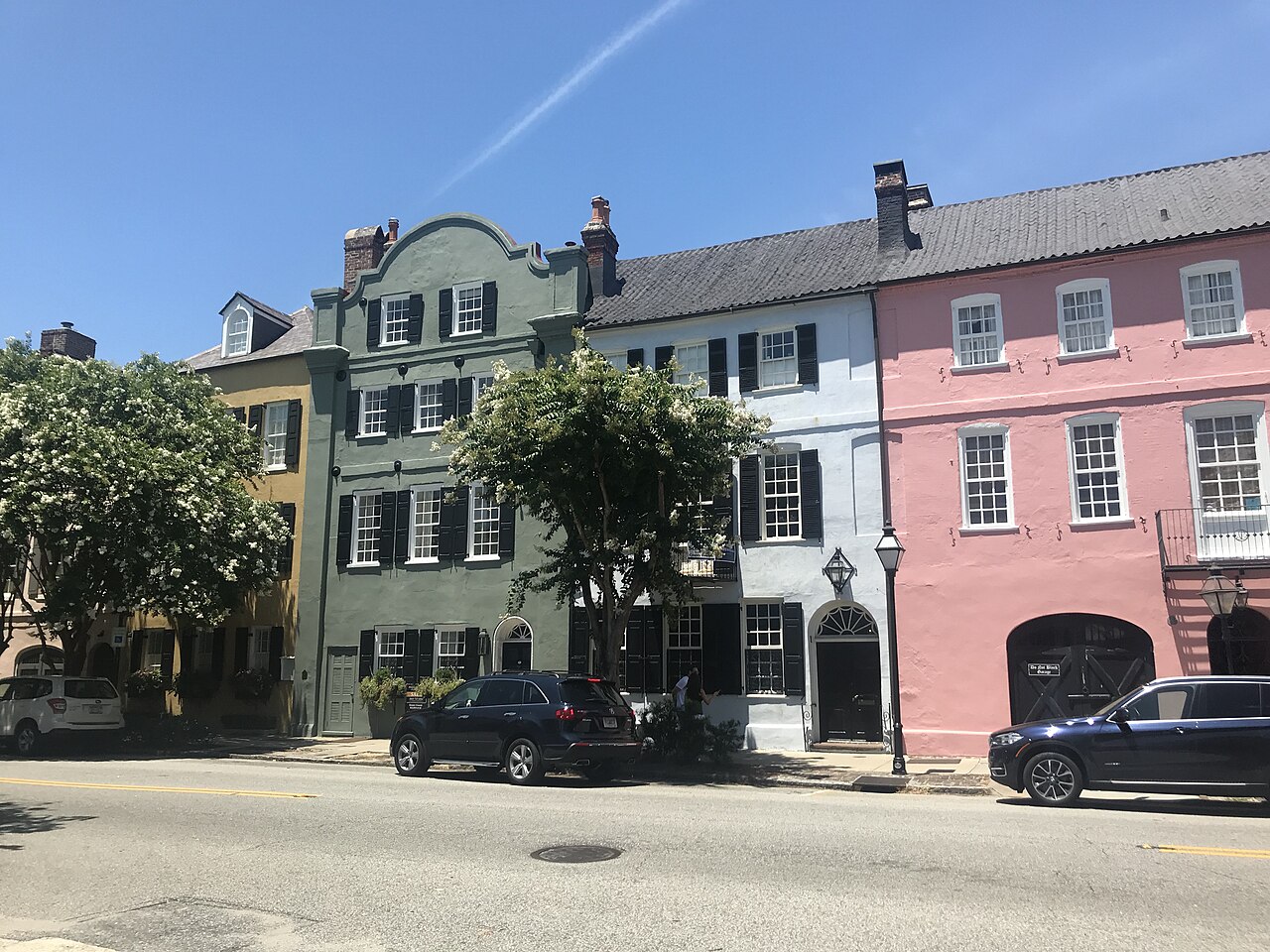A row of joined historic town houses painted green, grey and pink, with street trees and parked cars in front.