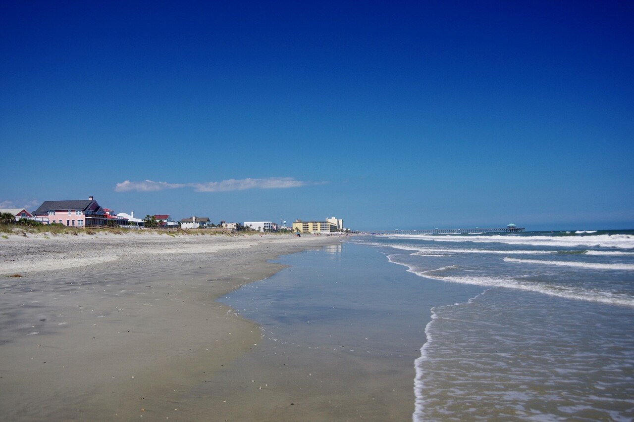 A wide, wet sand beach at low tide with a line of beach houses on the left and small waves on the right.