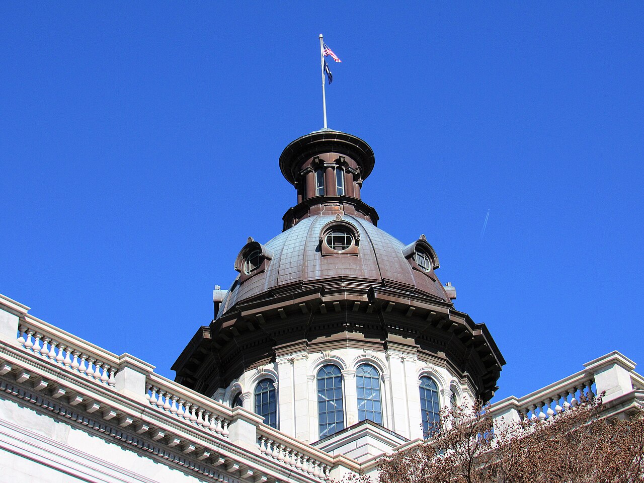 A copper-coloured dome with a ring of arched windows and a flagpole on top, seen from below against a blue sky.