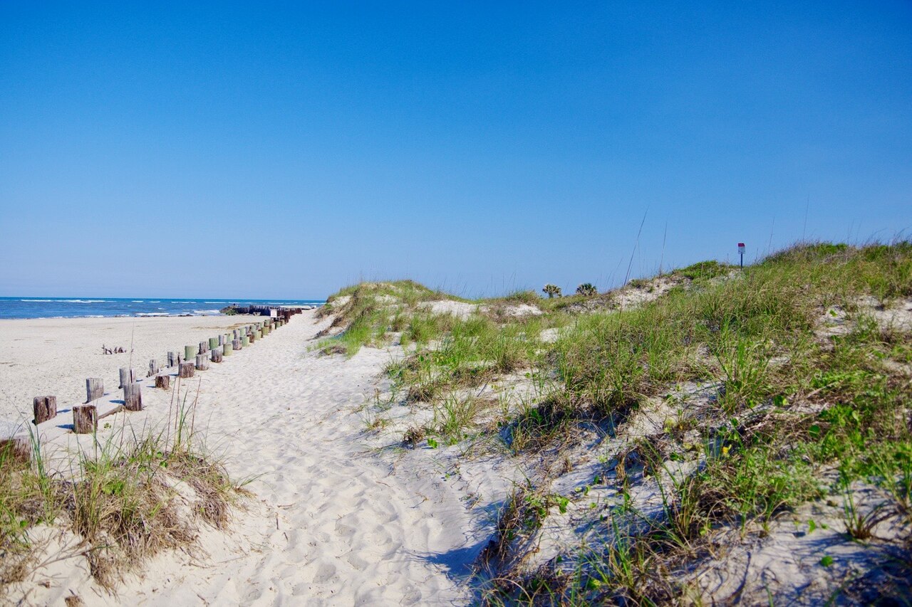 A sandy path between grass-covered dunes leading to the sea, with a line of wooden posts on the beach.