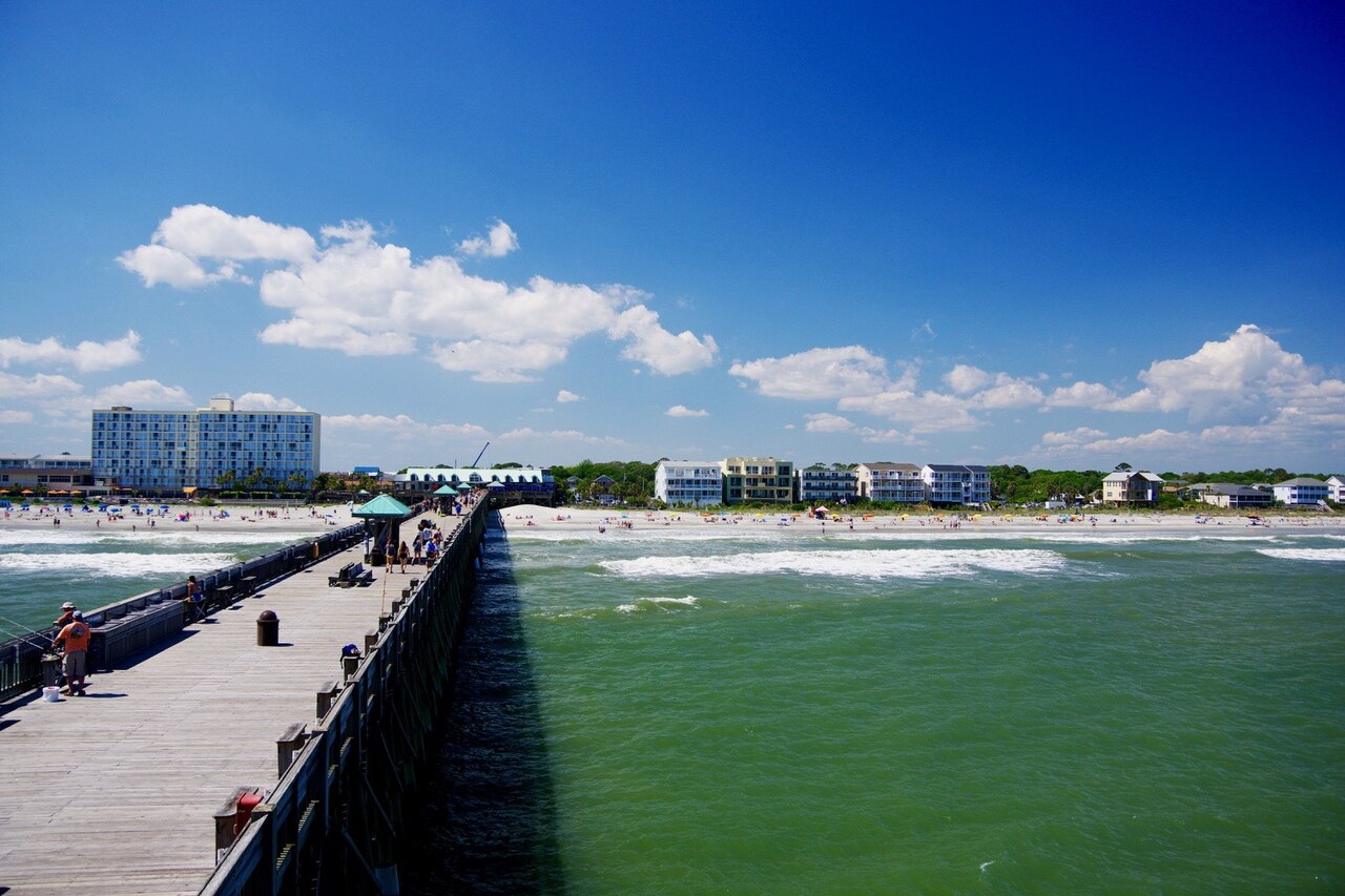 A long wooden pier running toward a shoreline of hotels and beach houses, with green water and white clouds.
