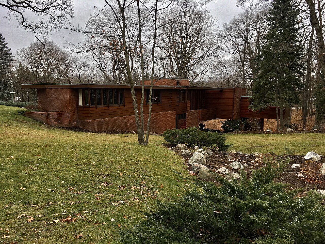 A long, low brick house with a flat overhanging roof on a grassy slope, with a rock-lined stream bed and tall trees.