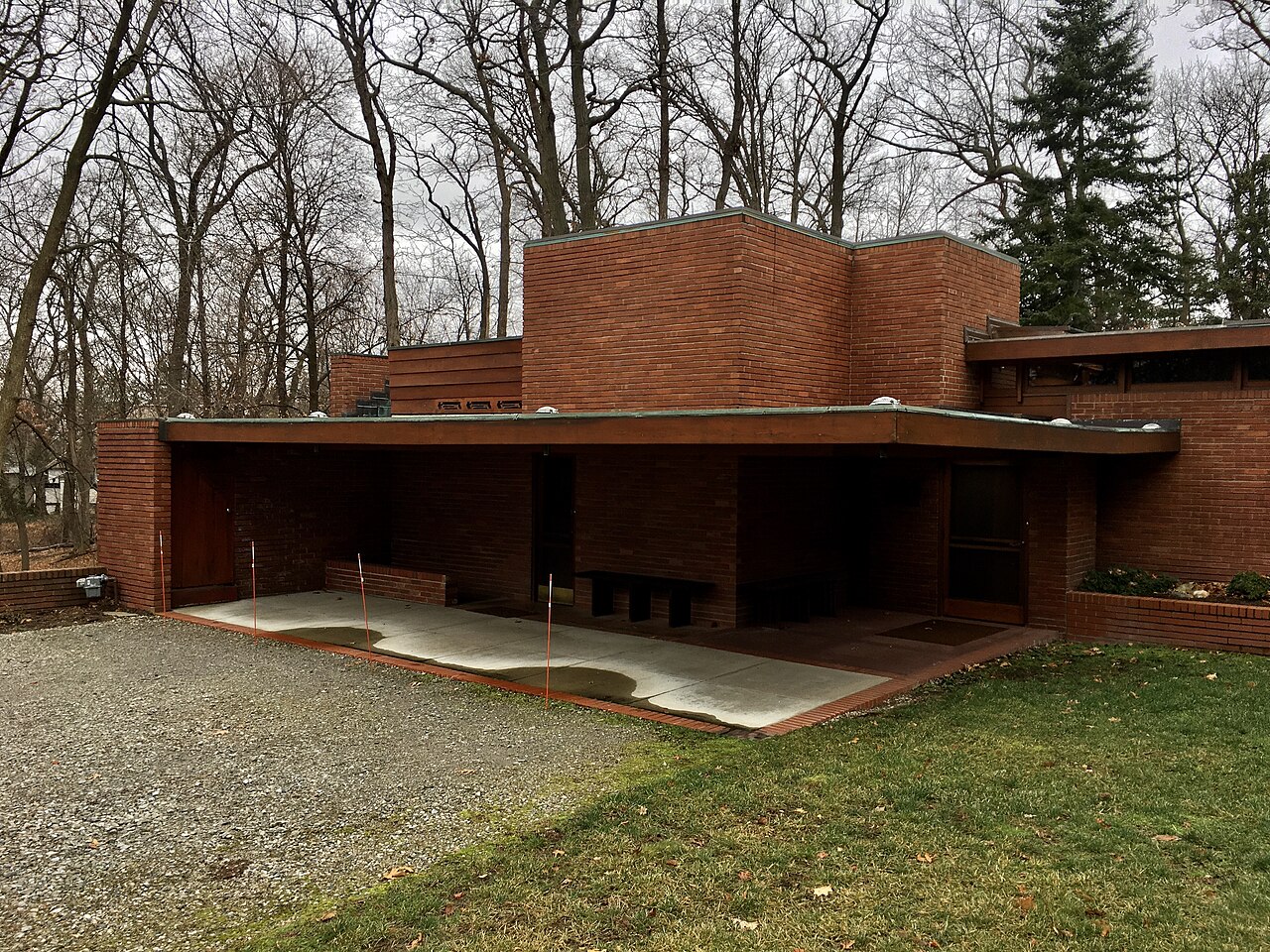 A low red brick house with a flat roof and a deep open loggia, set on a gravel drive among bare winter trees.