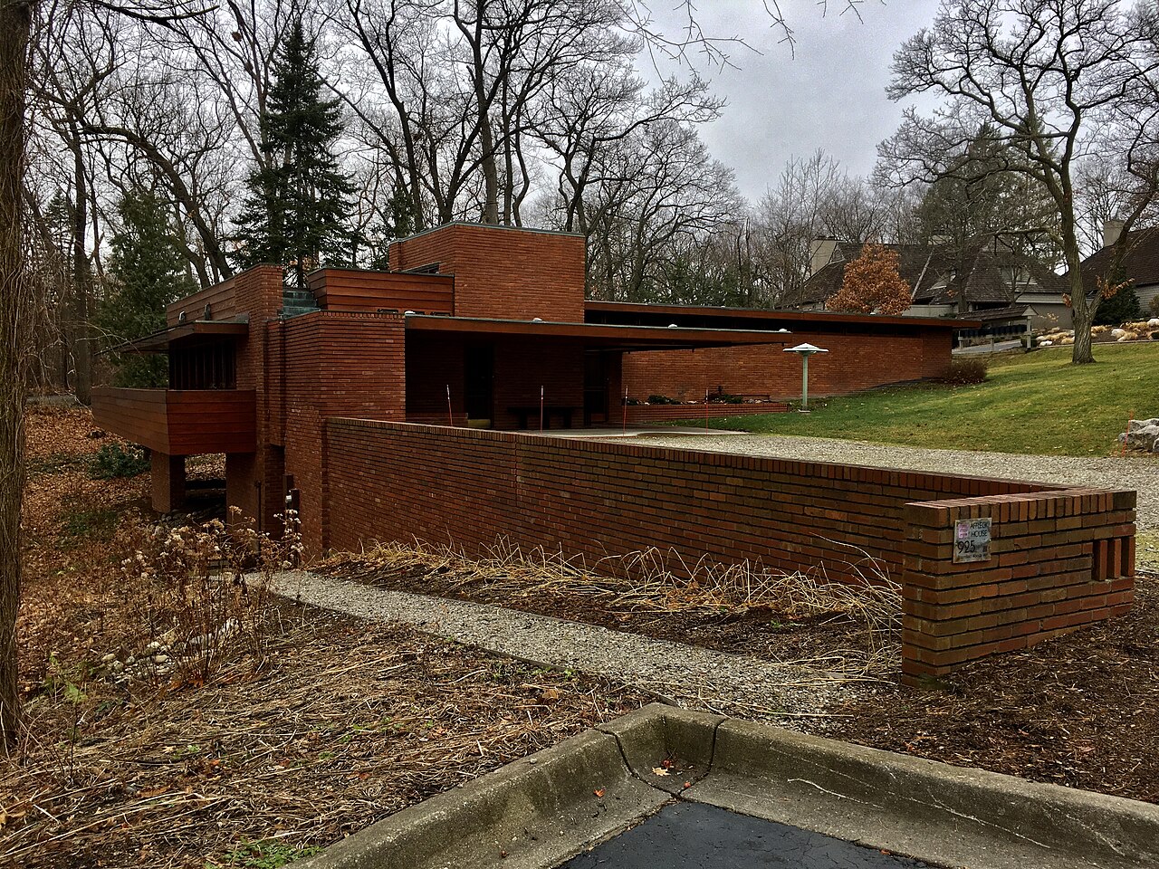 A low brick garden wall leading to a flat-roofed brick house with a cantilevered wing, among bare trees.