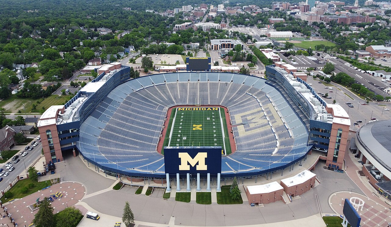 An aerial view of a huge empty football stadium bowl with a block M at midfield, and a city spreading out behind it.