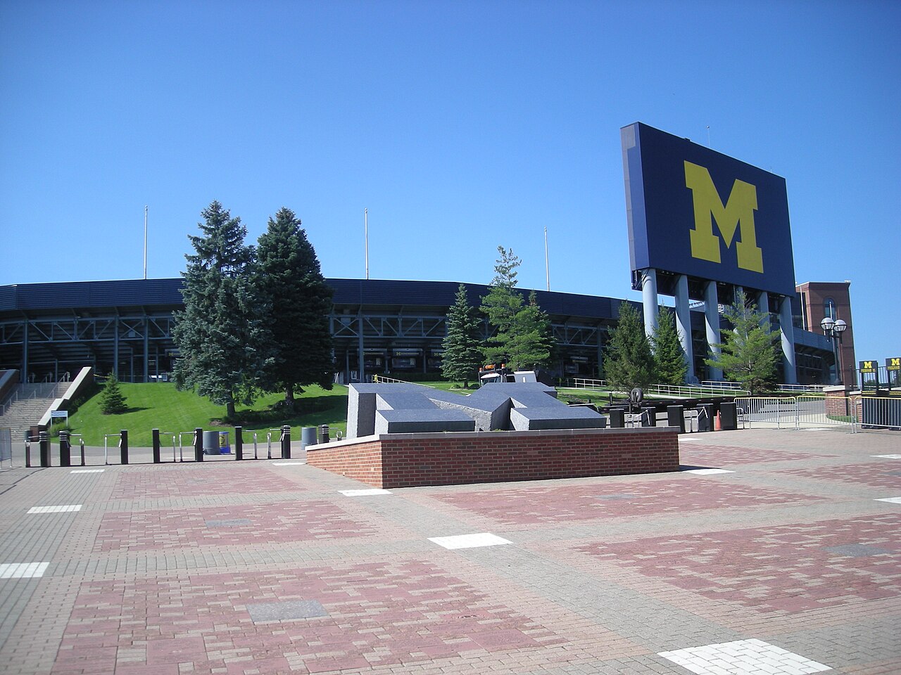 A brick plaza and low stadium wall with a tall blue scoreboard carrying a yellow block M, under a clear sky.