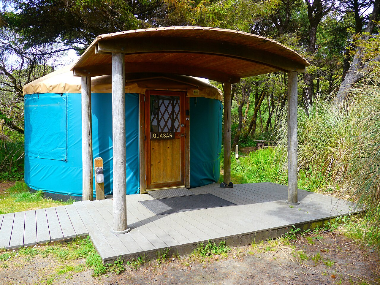 A round blue fabric yurt with a wooden door and a covered wooden porch, reached by a boardwalk through coastal trees.