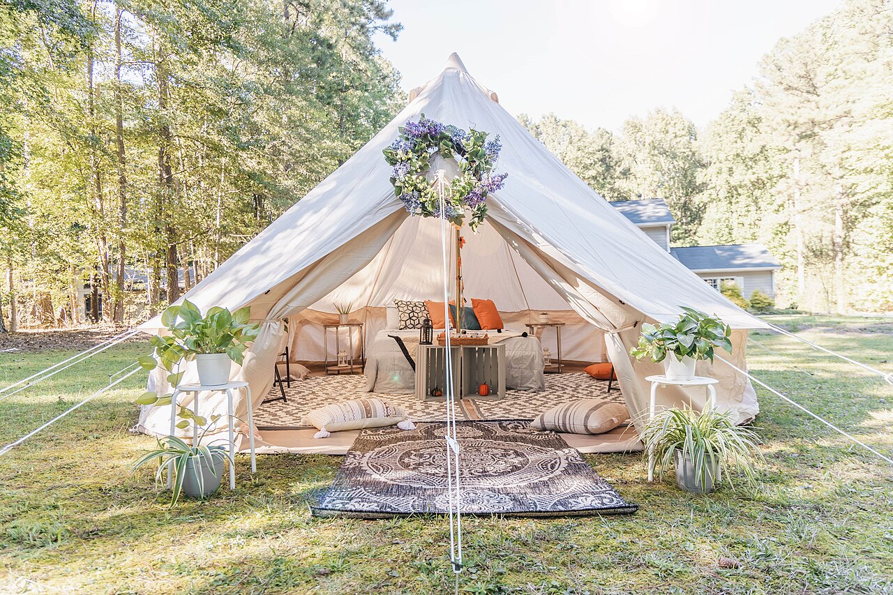 A cream canvas bell tent with its front rolled open, showing rugs, cushions and a low bed, on a lawn edged with pine trees.