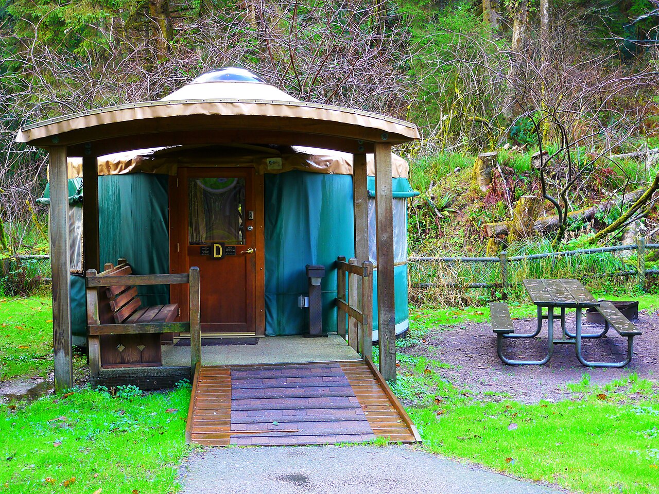 A round green fabric yurt with a wooden porch and ramp, a picnic table beside it, backed by dense forest.