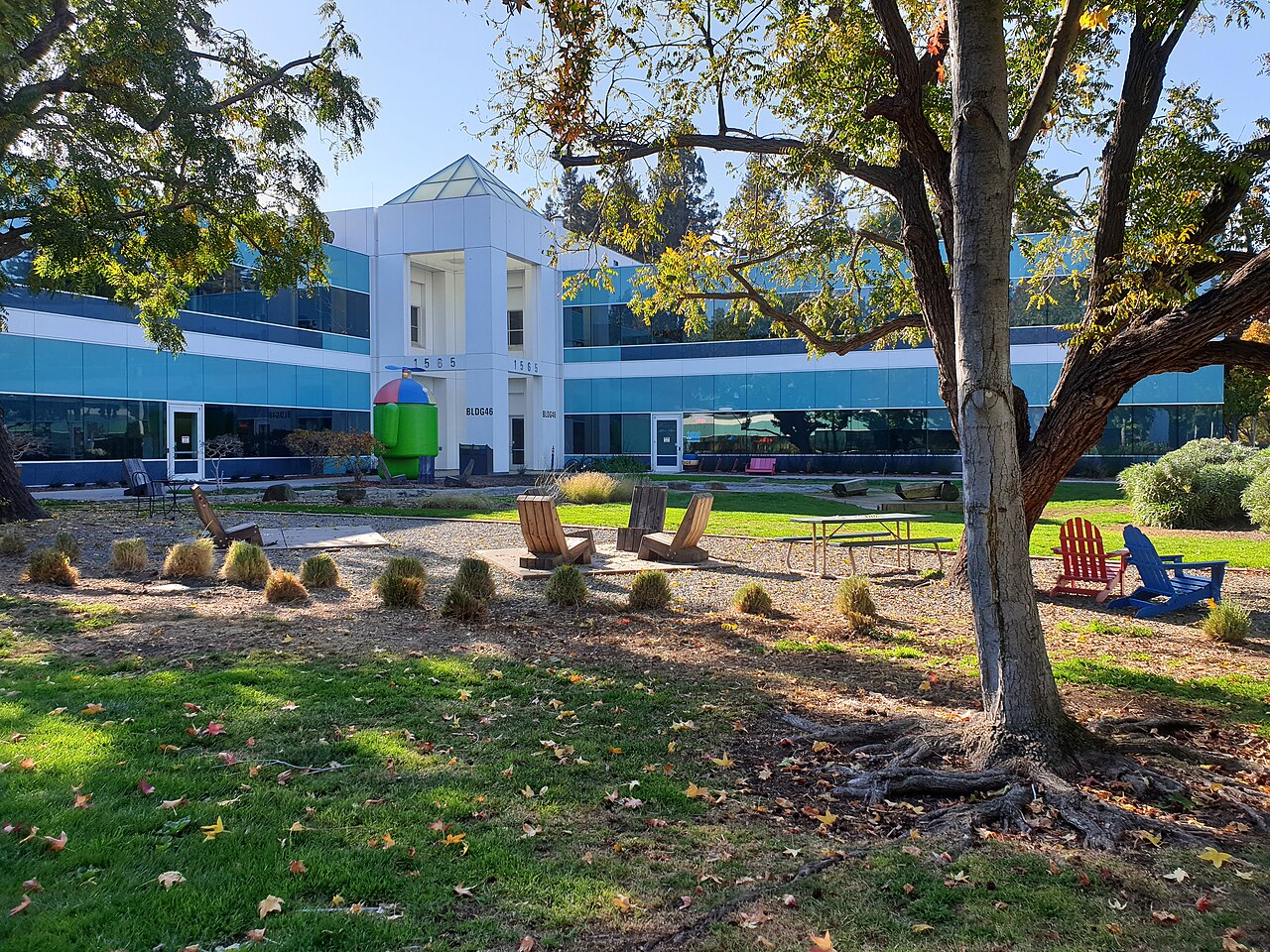 Low glass office buildings behind a lawn with garden chairs and a large tree in the foreground.