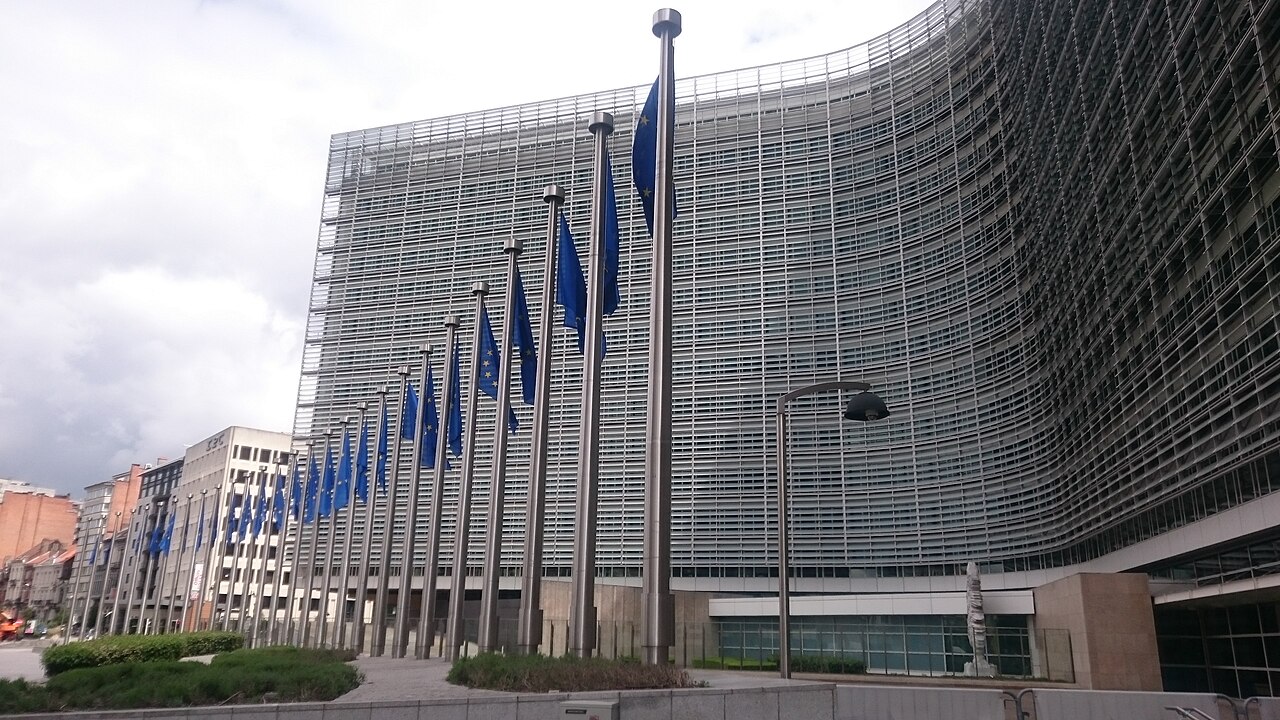 A curved glass and steel office building with a row of blue European Union flags on poles in front.