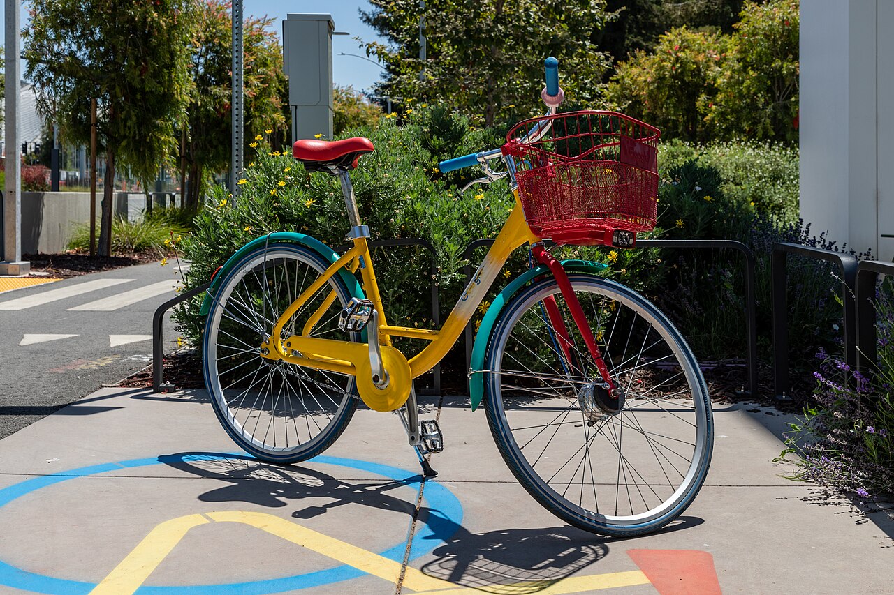 A yellow, green, blue and red bicycle with a front basket parked on a path beside shrubs.