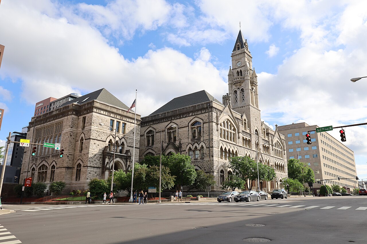 A grey stone Victorian Gothic building with a tall clock tower on a city street corner under a cloudy sky.