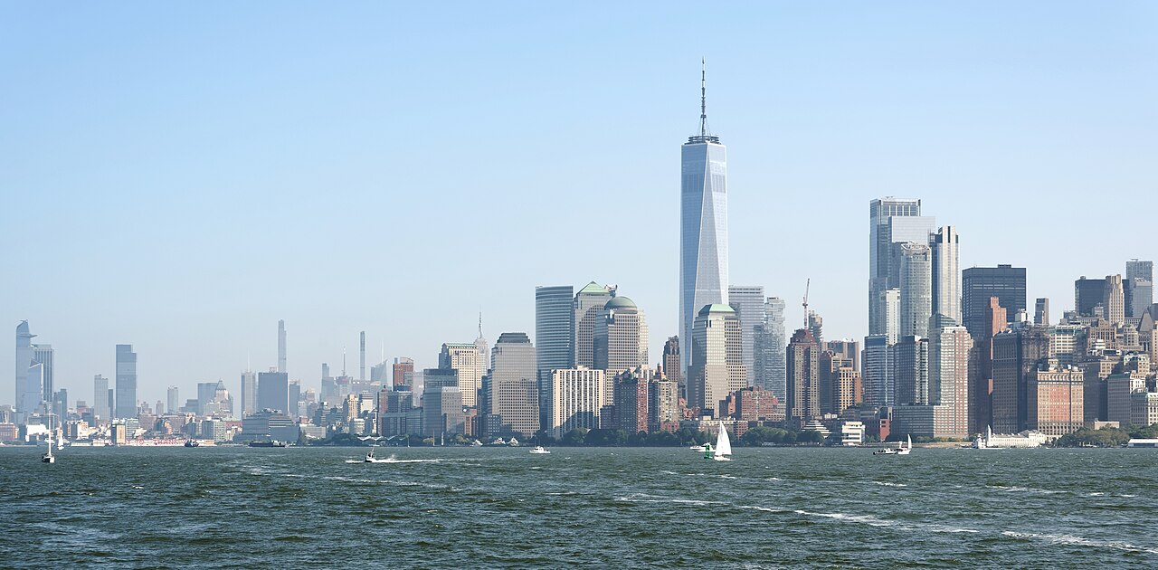 A cluster of skyscrapers with one tall spire rising above the rest, seen across open water under a clear sky.