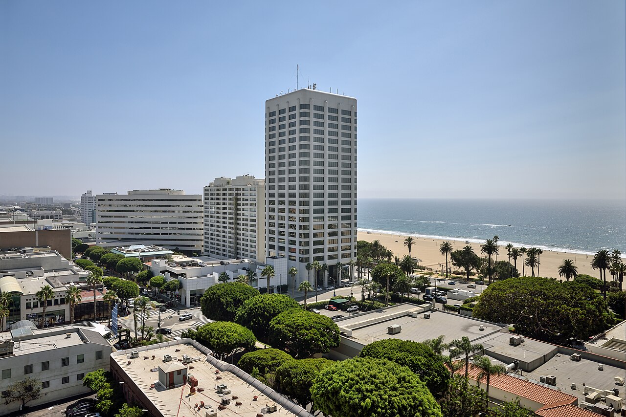A white office tower rising above low rooftops and trees, with a beach and the ocean behind it.